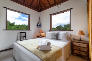 Quarto com cama, duas mesas de cabeceira, uma cadeira e janelas amplas com vista para o verde.