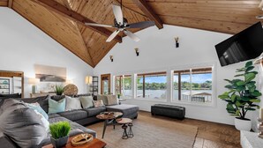 Ein geräumiges Wohnzimmer mit hoher Decke, großen Fenstern, einem grauen Sofa und einem Deckenventilator aus Holz.
