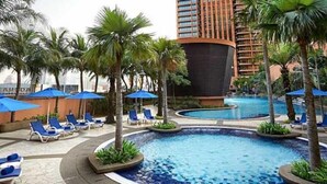 A hotel pool area with lounge chairs and umbrellas, surrounded by palm trees and a large building in the background.