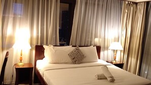 A hotel room with a large bed, two bedside lamps, and a view of the city at night through sheer curtains.