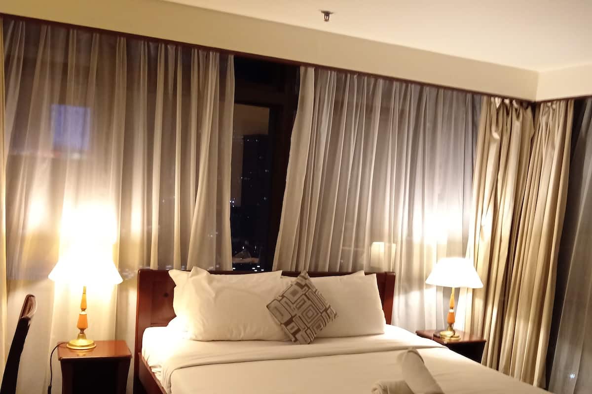 A hotel room with a large bed, two bedside lamps, and a view of the city at night through sheer curtains.