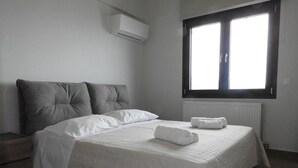 A neatly made bed with white bedding and a rolled towel, a grey upholstered headboard, a window with blinds, and an air conditioning unit on the wall. - Elea Sea View Apartment in Ag. Nikolaos (Agios Nikolaos)