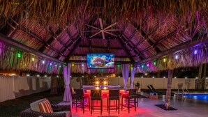An outdoor bar area with a thatched roof, a pool, and a TV screen.