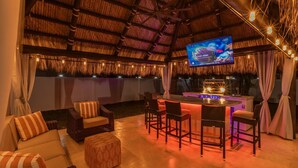 An outdoor bar area with a thatched roof, wicker furniture, and a flat-screen TV mounted on the ceiling.