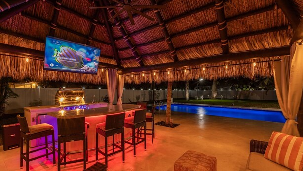 An outdoor bar area with a thatched roof, a large screen TV, and a pool in the background.