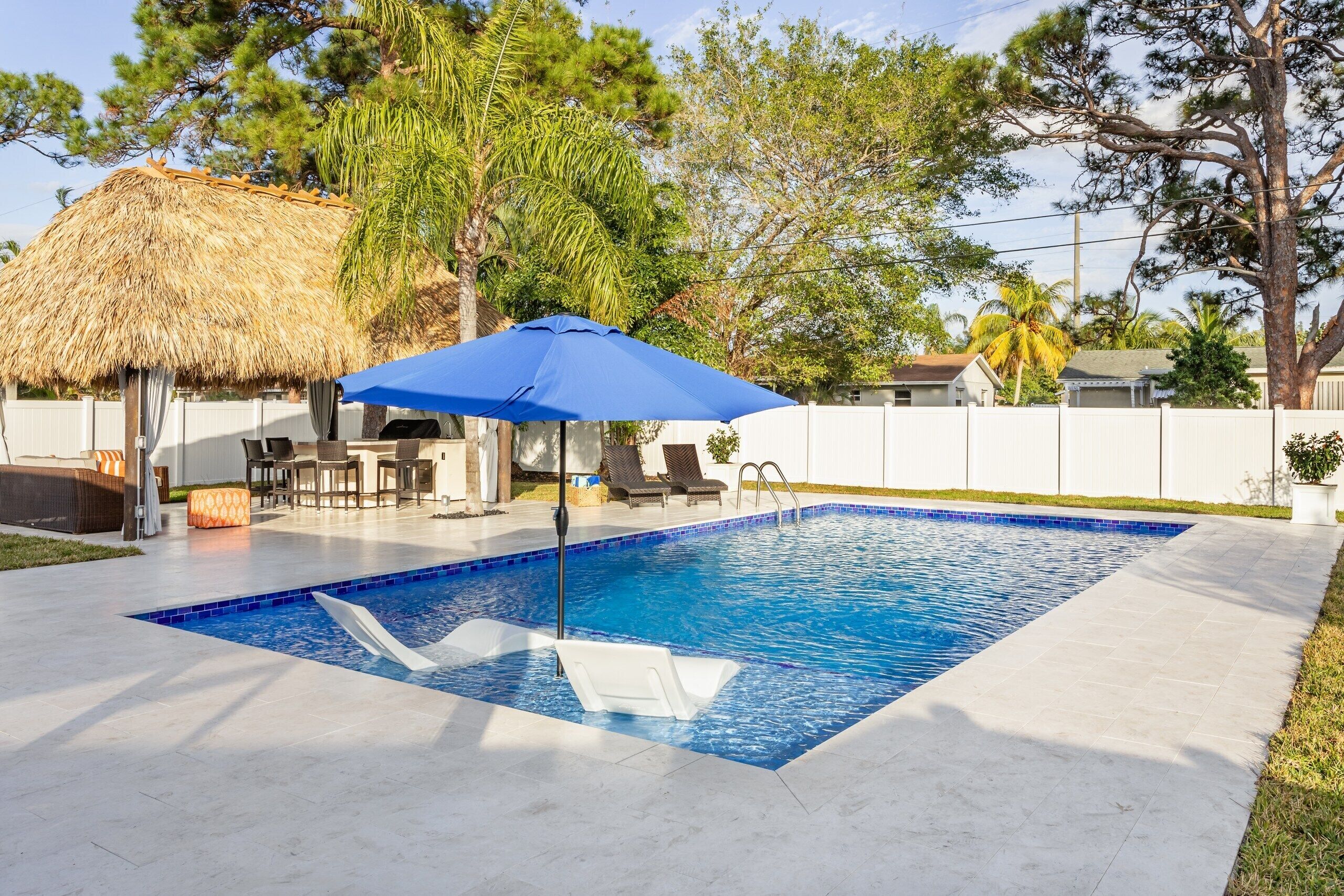 Una piscina con sillones de descanso y una sombrilla azul, rodeada por una valla blanca y árboles.
