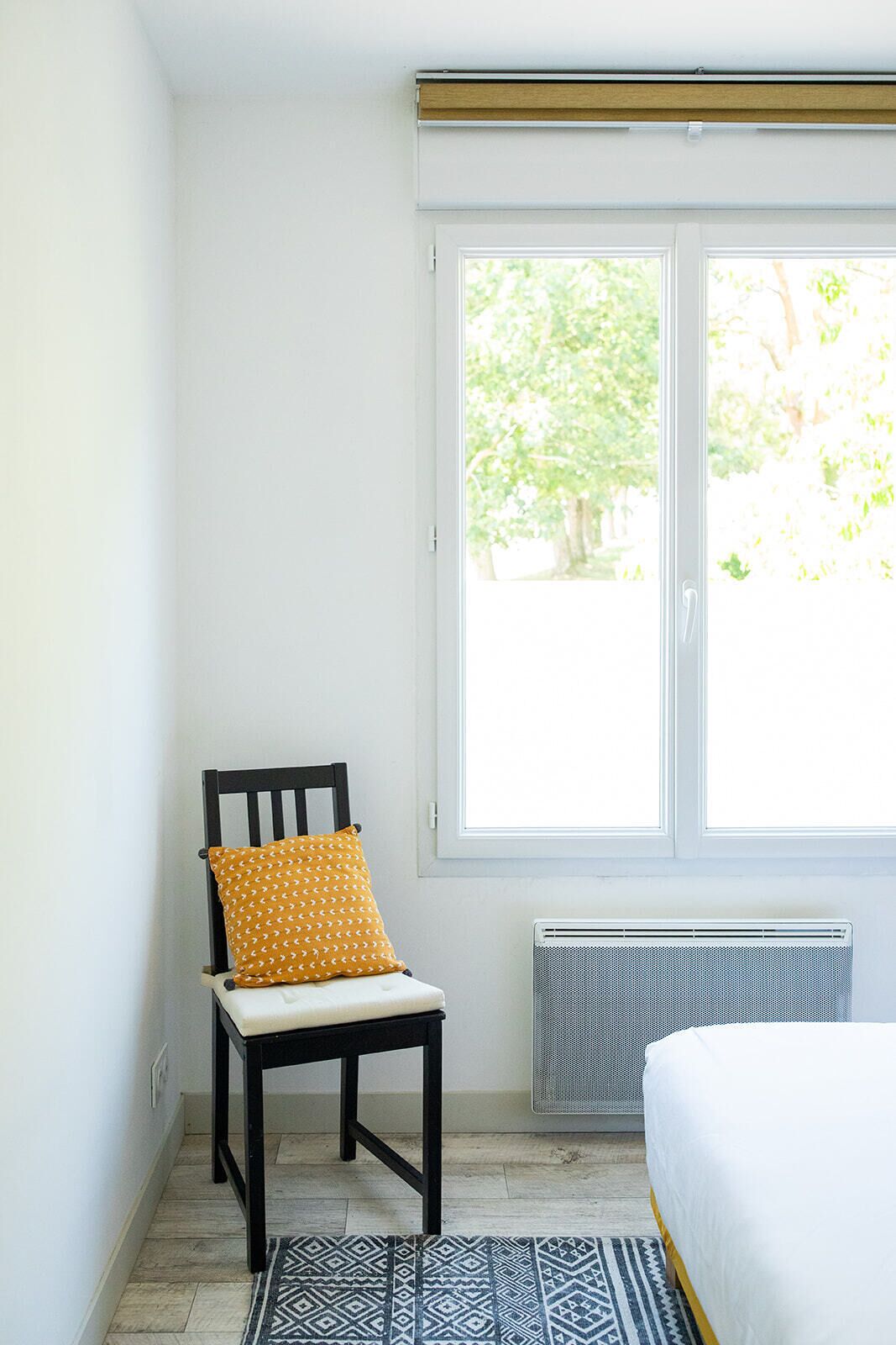 A bedroom with a bed, a chair with a patterned cushion, a radiator, and a window with a view of greenery.