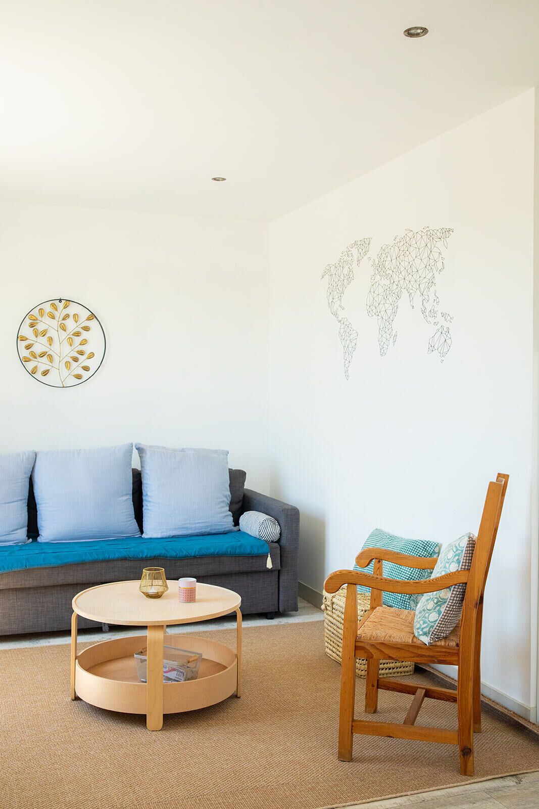Un salon avec un canapé gris, une table basse ronde en bois, un fauteuil en osier et une décoration murale représentant une carte du monde.