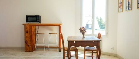 A small, open-plan living space with a wooden dining table, two bar stools, a kitchen area with a microwave, and a large window providing natural light.