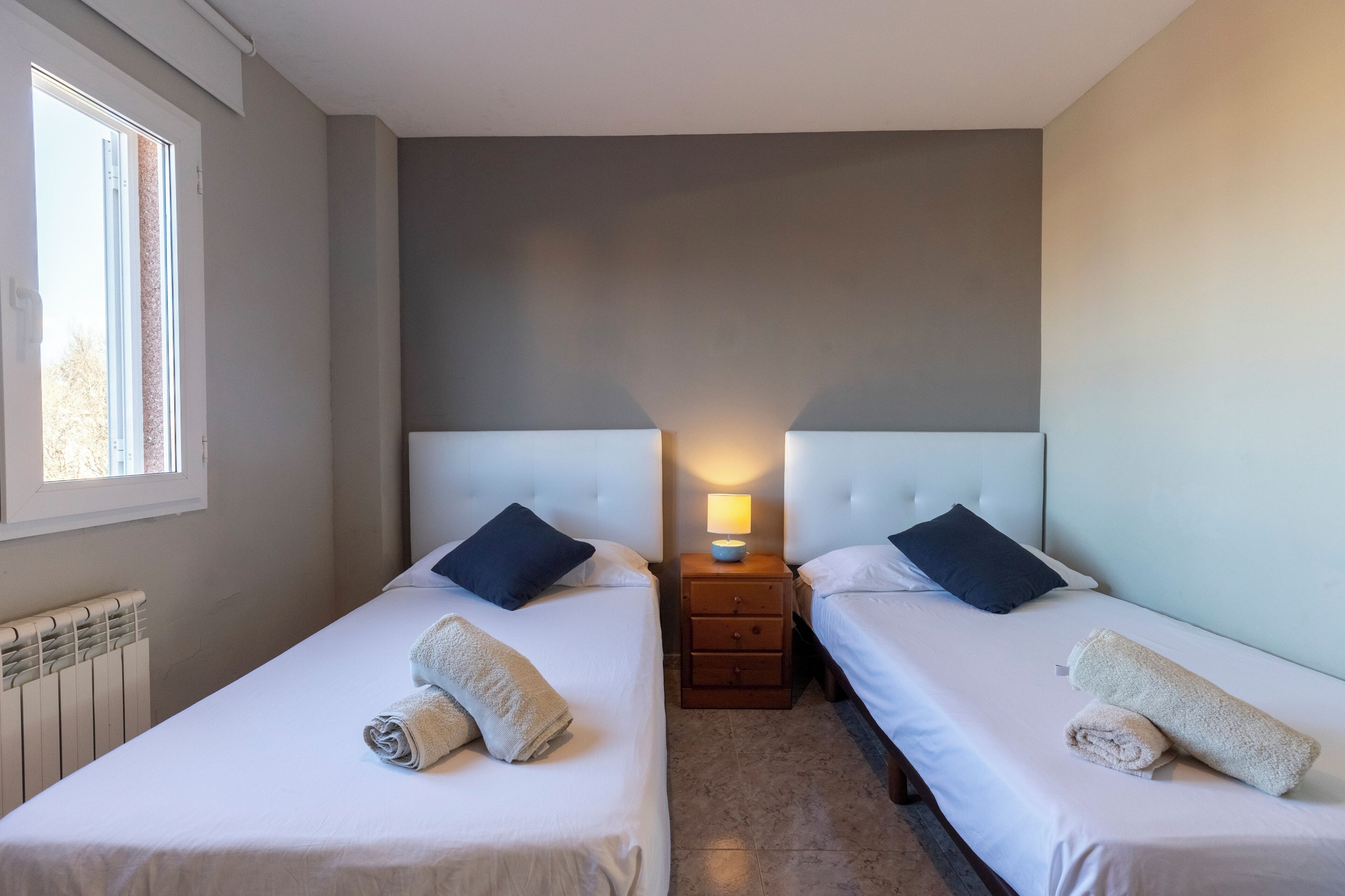 A hotel room with two single beds, each with a pillow, a bedside table with a lamp, and rolled towels on the beds.