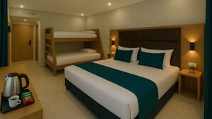 Une chambre d’hôtel avec un lit superposé, un lit simple, une table de chevet avec une bouilloire et du café, et une petite table de nuit avec une lampe.