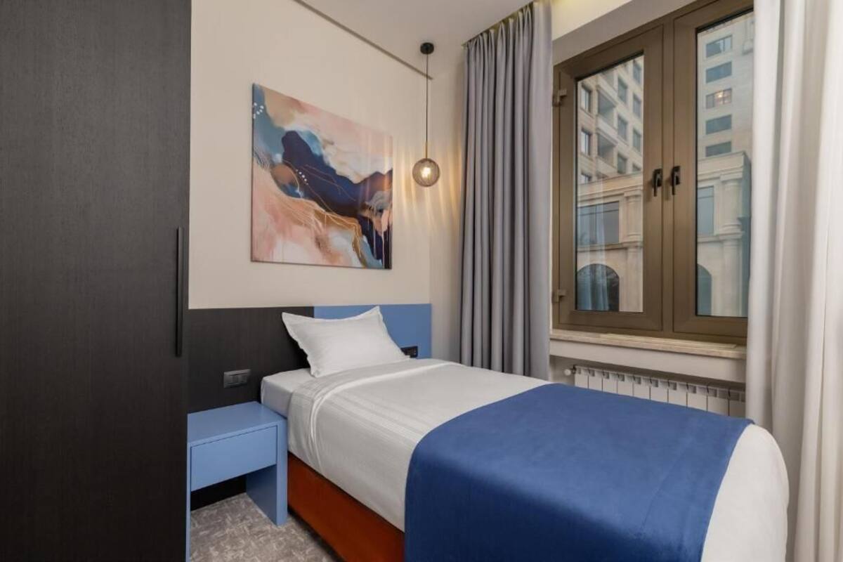 Hotellihuone, jossa on sänky, yöpöytä, taulu seinällä, ikkuna verhoilla ja näkymä ulkona oleviin rakennuksiin.