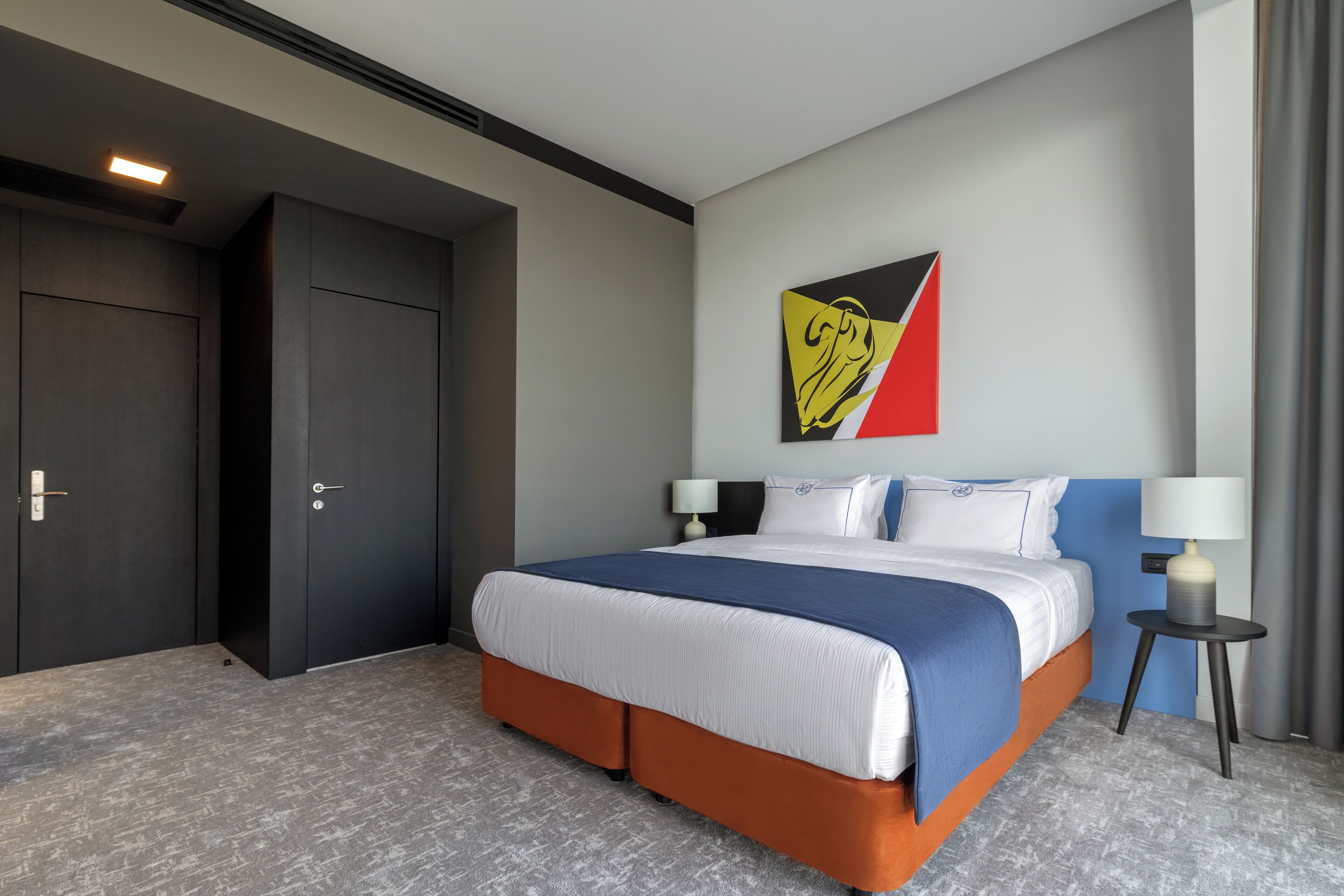 A modern hotel room with a large bed, a nightstand, a wall art piece, and a door.