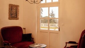 A room with a red armchair, a wooden chair, a small table, a chandelier, and a window with a view of trees and a white fence.