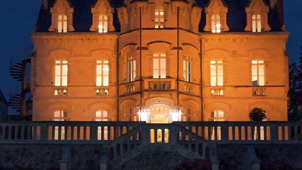 A large, illuminated building with a symmetrical facade, multiple windows, and a central entrance with steps leading up to it.