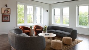 A spacious living room with a grey sectional sofa, two armchairs, a coffee table, and a rug. Large windows provide natural light and views of greenery outside. - Historic Rooftop Steps to DT Shops & Dining Unit 3 (Tuscaloosa)