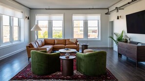 A modern living room with a sectional sofa, two green armchairs, a coffee table, and a TV on a wooden cabinet. Large windows provide natural light and views of buildings outside. - Historic Rooftop Steps to DT Shops & Dining Unit 5 (Tuscaloosa)