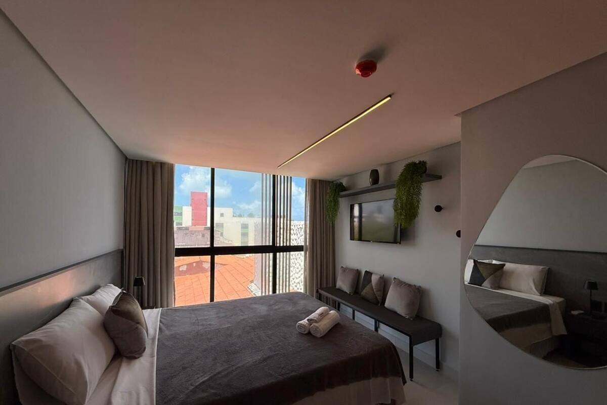 A modern hotel room with a large bed, a seating area, a TV, and a view of the city.