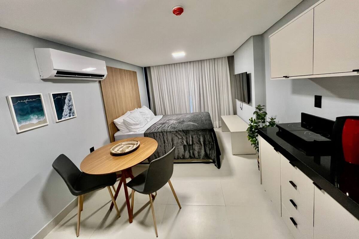 A compact hotel room with a bed, a small dining table with chairs, a kitchenette, a TV, and an air conditioning unit.