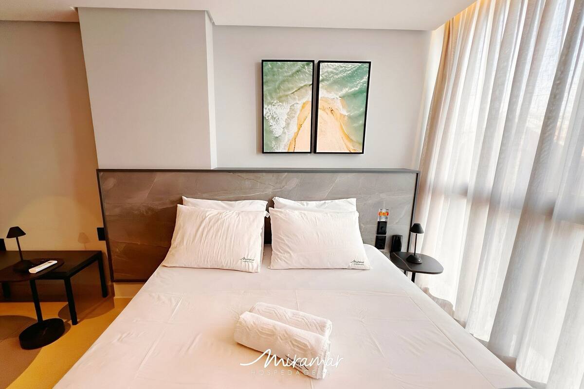 A hotel room with a large bed, two pillows, a bedside table with a lamp, a remote control, and a wall-mounted artwork.