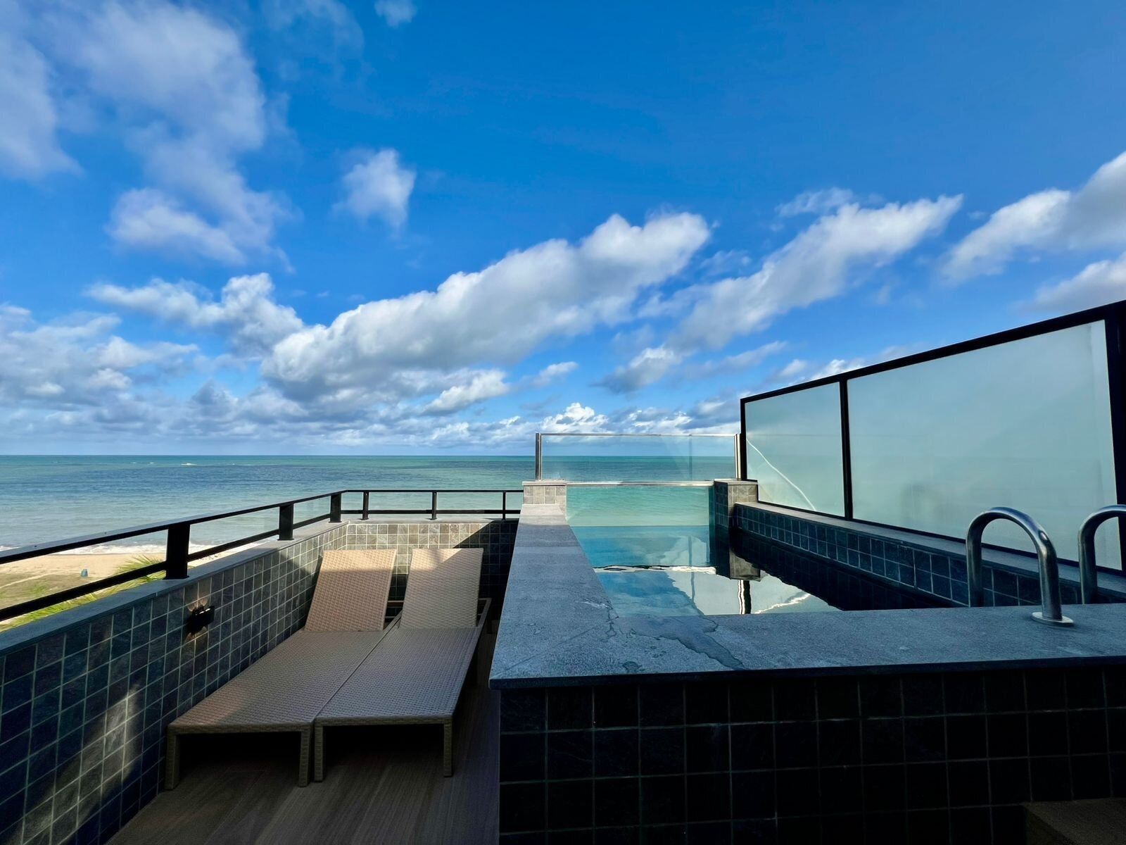 A pool area with a view of the ocean, a glass barrier, and a tiled floor.