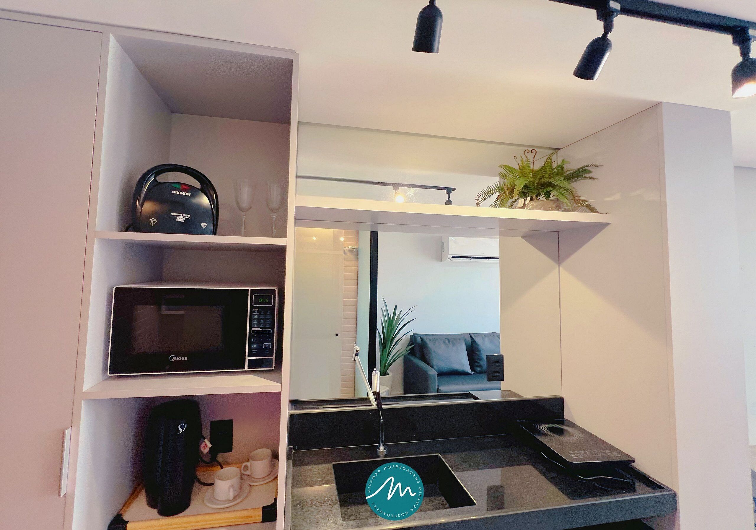 Eine moderne Küche mit Mikrowelle, Wasserkocher und Kaffeemaschine. Auf der Arbeitsfläche steht eine kleine Pflanze, und es gibt eine Sitzecke mit Sofa und Couchtisch.
