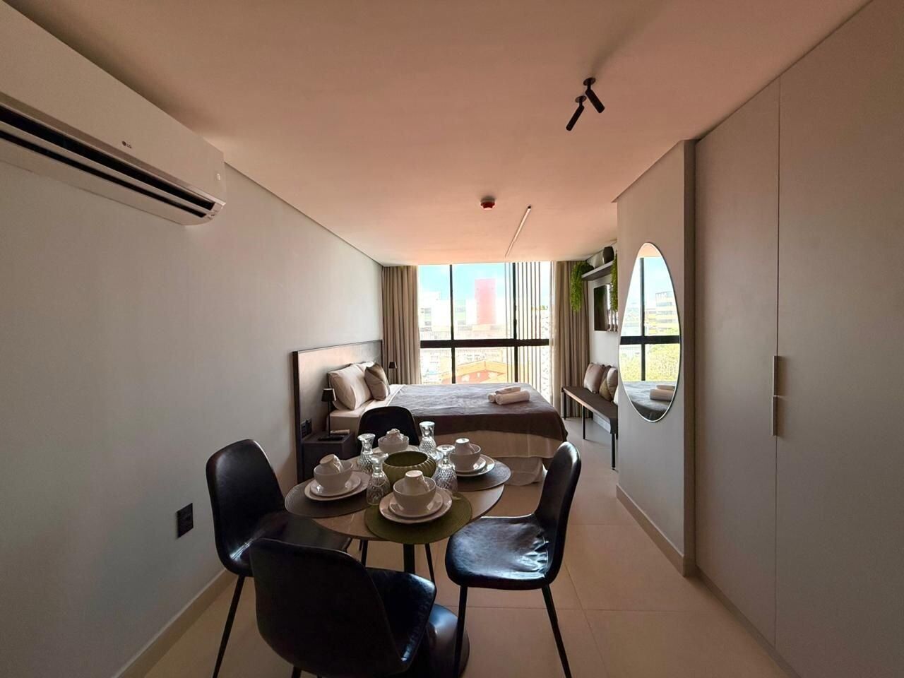 A hotel room with a bed, a dining area with a table set for two, a large window with a view, and a wardrobe.