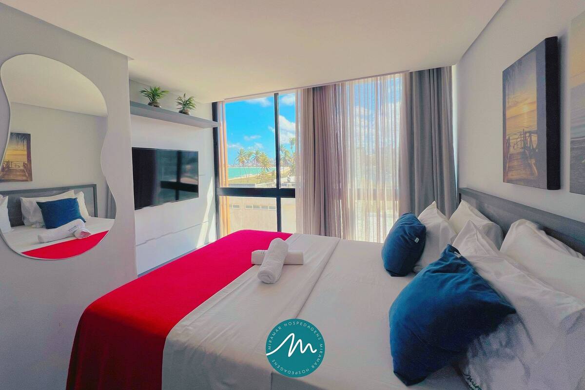 A hotel room with a large bed, a TV, a window with a view of the beach, and a red accent wall.