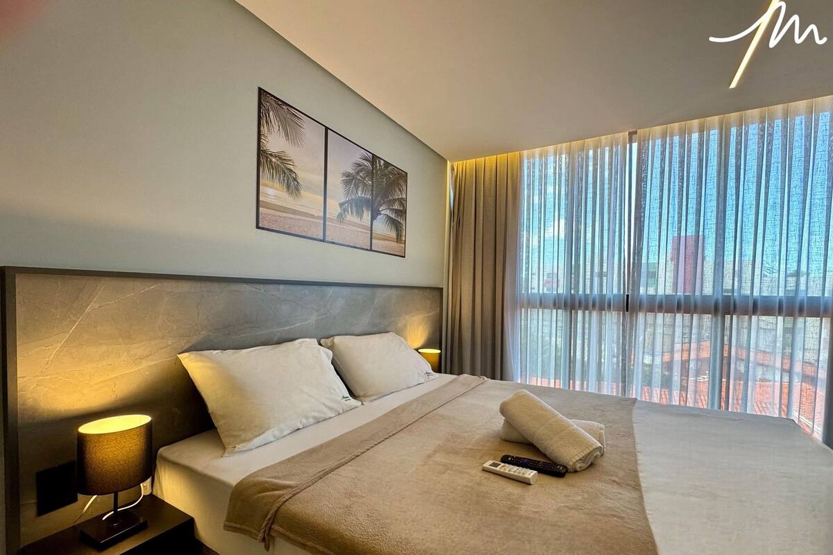 A hotel room with a large bed, two bedside tables with lamps, a headboard, and a view of the city through sheer curtains.