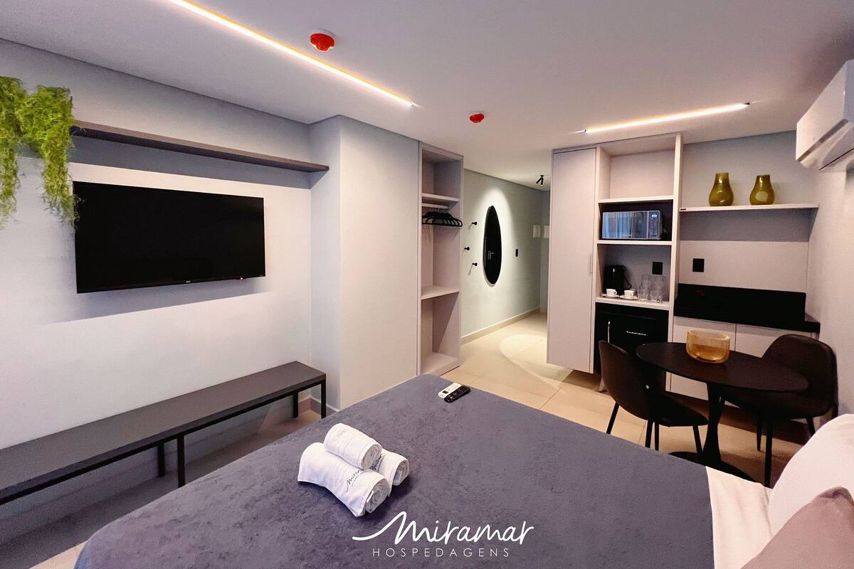 A modern hotel room with a large bed, a bench, a TV mounted on the wall, a desk with chairs, and a small dining area.