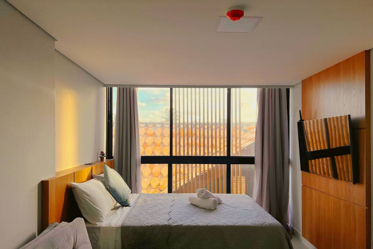 A hotel room with a large window, a bed with pillows, a wooden headboard, and a mounted TV.