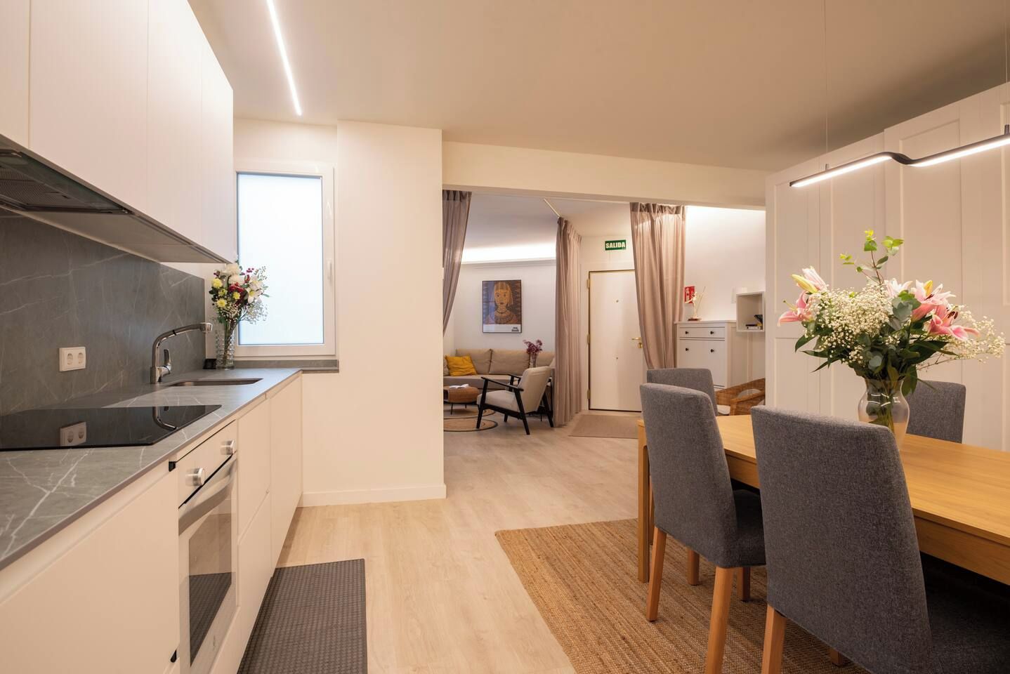 Una cocina moderna con estufa integrada, un comedor con mesa de madera y sillas grises, y una sala de estar con sofá y un cuadro en la pared.