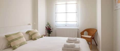 A neatly made bed with pillows and folded towels, a wicker chair, and a window with blinds.