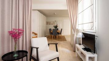 Een moderne woonkamer met een televisie, een eethoek op de achtergrond en een bloevas op een bijzettafeltje.