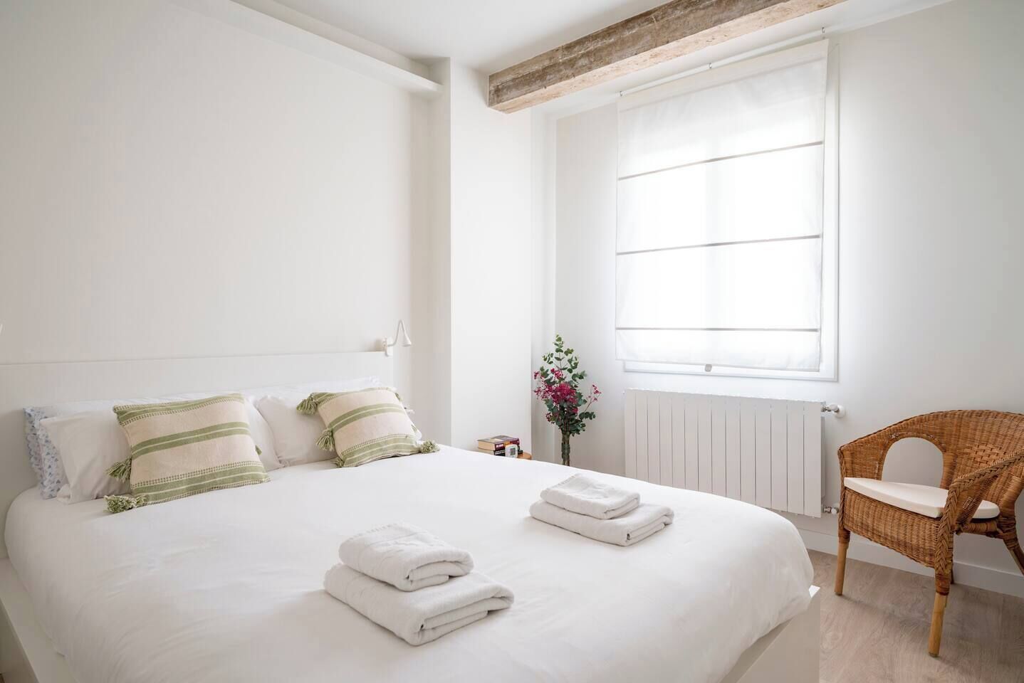 Ein ordentlich bezogenes Bett mit Kissen und gefalteten Handtüchern, ein Korbstuhl und ein Fenster mit Jalousien.