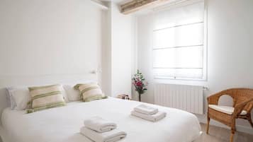 Ein ordentlich bezogenes Bett mit Kissen und gefalteten Handtüchern, ein Korbstuhl und ein Fenster mit Jalousien.