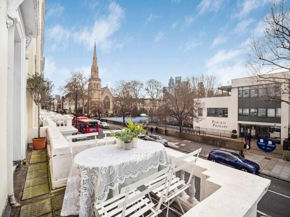 Un balcon avec une table et des chaises, donnant sur une rue où se trouvent une église et un bâtiment abritant une école primaire.