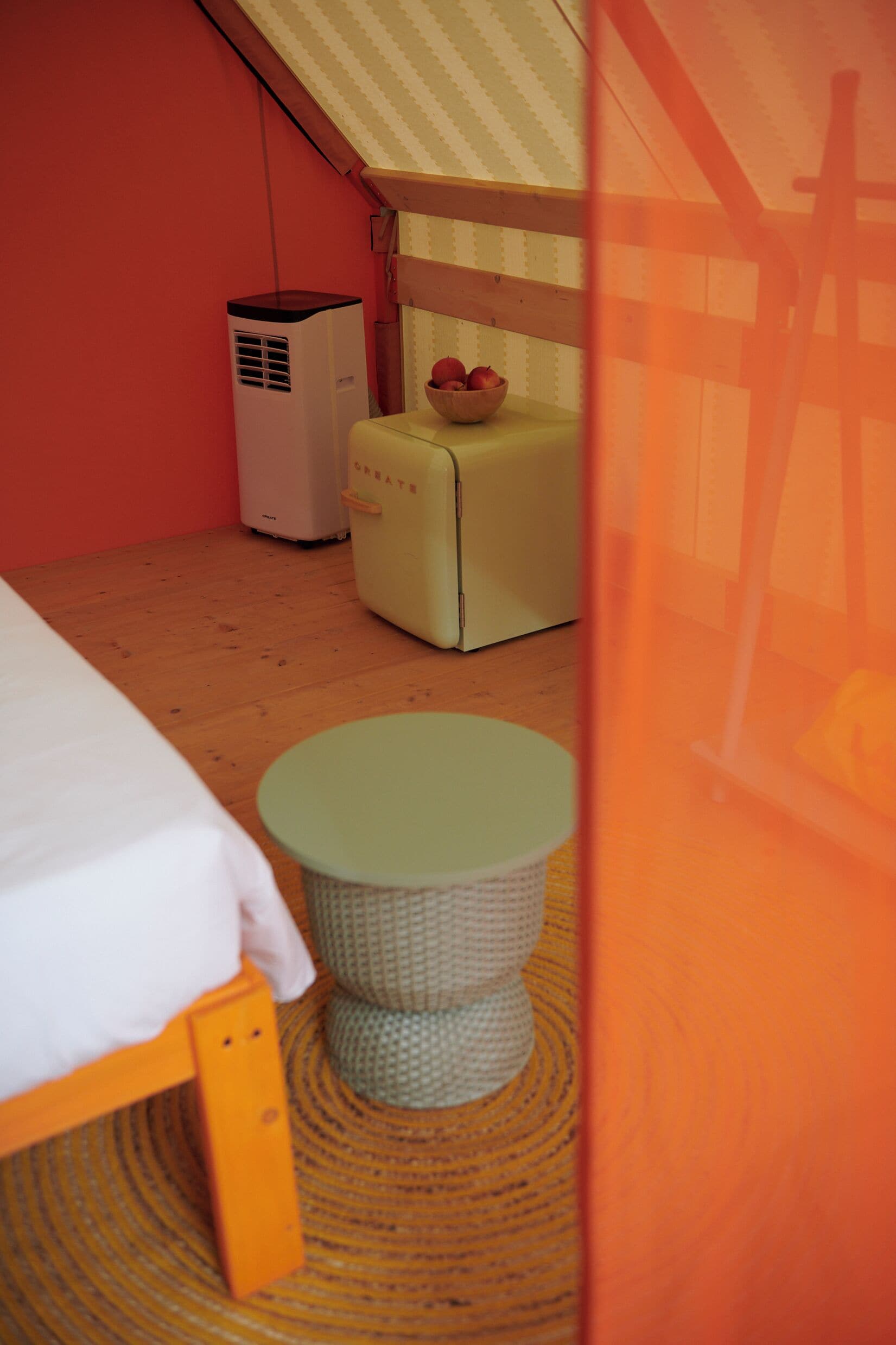 Een kleine, gezellige kamer met een bed, een krukje, een vintage koelkast en een raam met een gestreept gordijn.