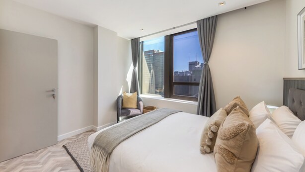 A modern hotel room with a large bed, a view of the city, and a window with curtains. - Designer Foreshore Apartment | No Loadshedding (Cape Town)