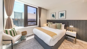 Een moderne hotelkamer met een groot bed, een nachtkastje met een lamp, een stoel, een raam met gordijnen en twee ingelijste foto’s aan de muur.