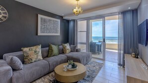 A modern living room with a grey sofa, a round coffee table, a chandelier, and a large window with a view of the ocean. - Lagoon Beach Escape with Breathtaking Views (Cape Town)