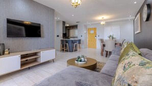 A modern living room with a flat-screen TV, a sofa, a coffee table, and a dining area with a kitchen in the background. - Lagoon Beach Escape with Breathtaking Views (Cape Town)