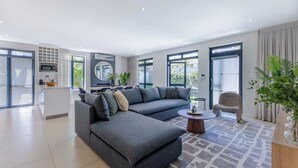 A modern living room with a grey sectional sofa, a round coffee table, and a grey armchair. There is a kitchen area with a microwave and a small island. Large windows provide natural light and views of the outdoors. - Three Bedroom Family Haven  Sea Point (Cape Town)