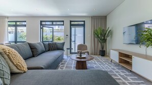 A modern living room with a grey sofa, a round coffee table, a TV mounted on the wall, and a potted plant. - Three Bedroom Family Haven  Sea Point (Cape Town)