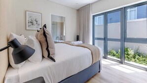 A modern bedroom with a large bed, a bedside lamp, a mirror, and a sliding glass door leading to a balcony with greenery. - Three Bedroom Family Haven  Sea Point (Cape Town)