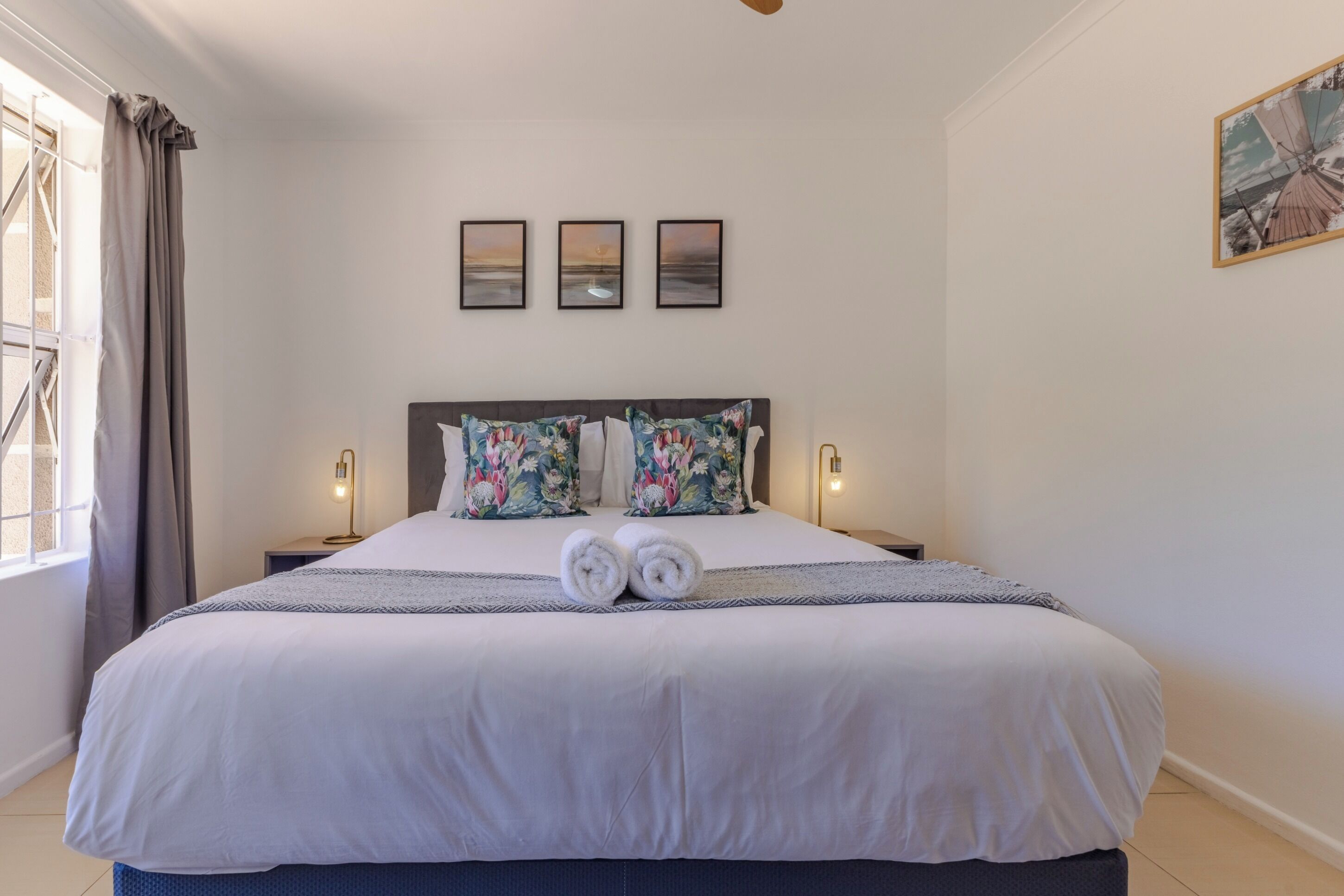 Ein ordentlich gemachtes Bett mit weißen Leinen, zwei Kissen mit Blumenmuster und zwei aufgerollte Handtücher auf dem Bett. Darüber an der Wand hängen drei gerahmte Bilder.