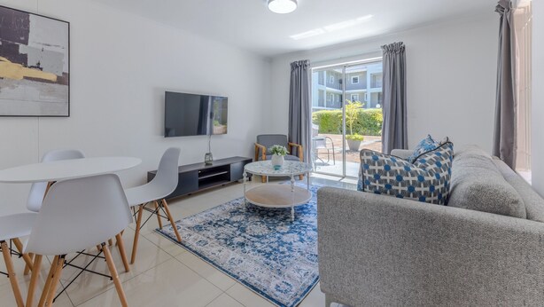 A modern living room with a sofa, a dining table with chairs, a TV mounted on the wall, and a large window with curtains. - 2-Bedroom Family Retreat in Gardens | Pool (Cape Town)