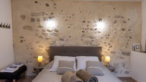 Un dormitorio con pared de piedra, una cama con ropa de cama blanca y gris, dos lámparas de noche y una mesita de noche con una lámpara.