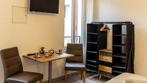 Un pequeño espacio de estar con una mesa de madera, dos sillas, un televisor montado en la pared, un estante negro y una ventana con vistas.