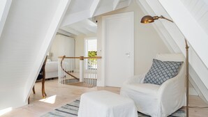 A cozy attic bedroom with a white chair, a small ottoman, and a floor lamp. A staircase leads to another room, and a door is visible in the background.